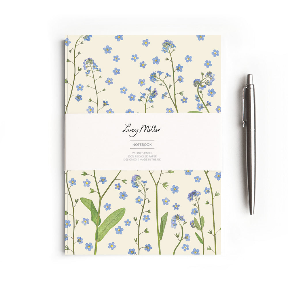 Notebook with pressed forget-me-not floral design with a belly band featuring 'Lucy Miller' branding. On a white background. Styled with a pen to show scale.