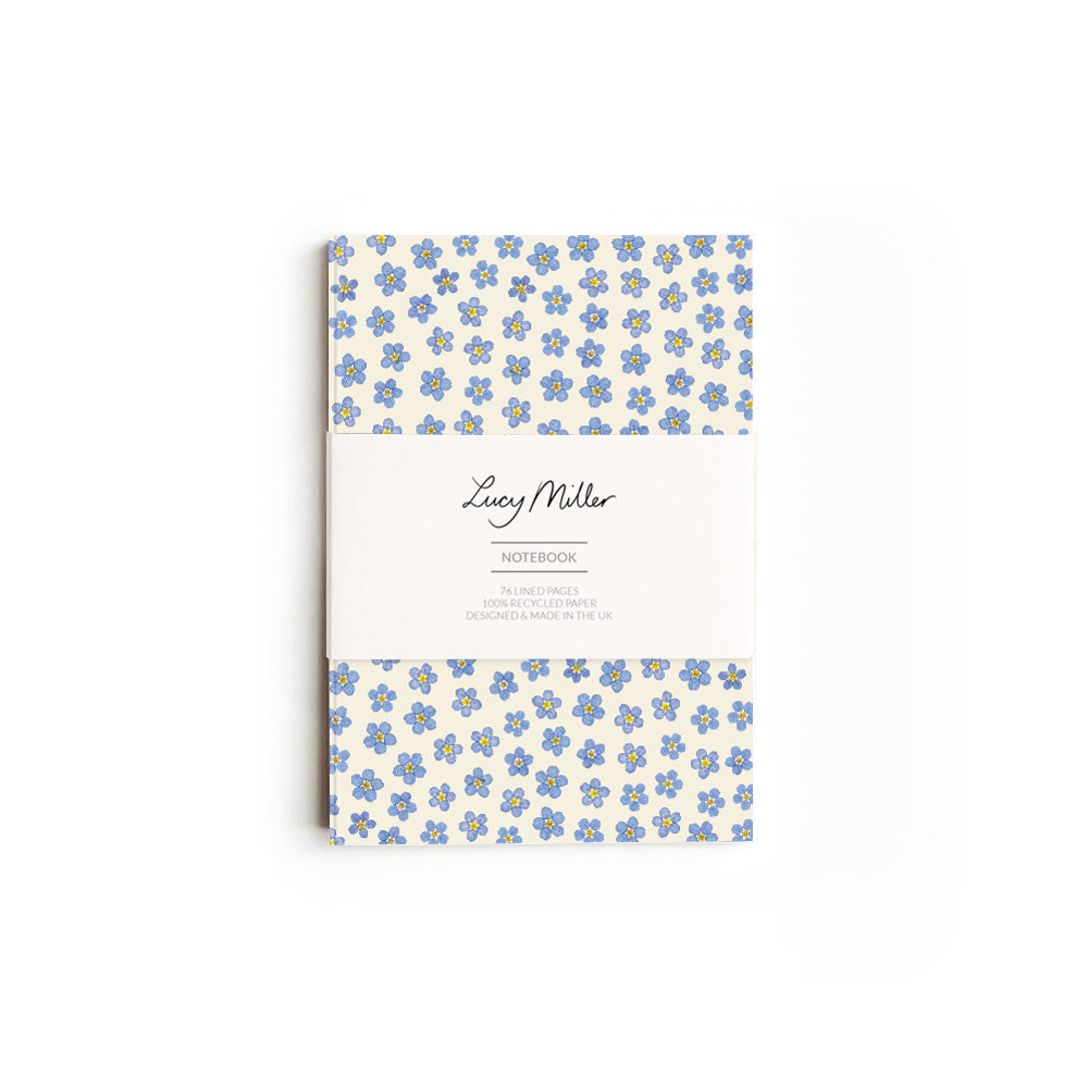 Notebook with pressed forget-me-not flowers with 'Lucy Miller' branded belly band. On a white background.