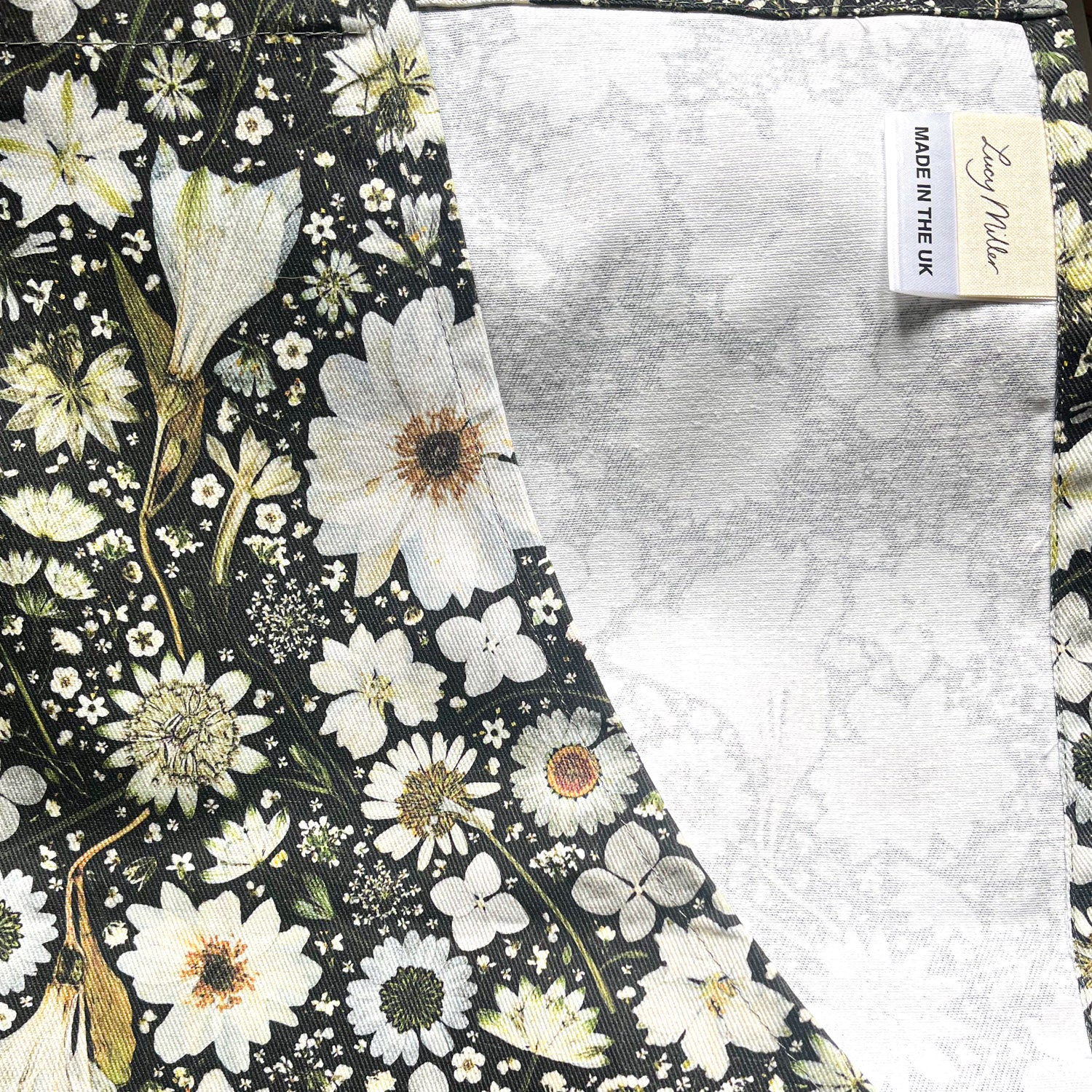A close-up Lucy Miller's floral apron. Featuring a design of white flowers on a black background. Flowers include Anemone, Nigella, Daisy, Astrantia, Heather, Hydrangea and Queen Anne’s Lace.