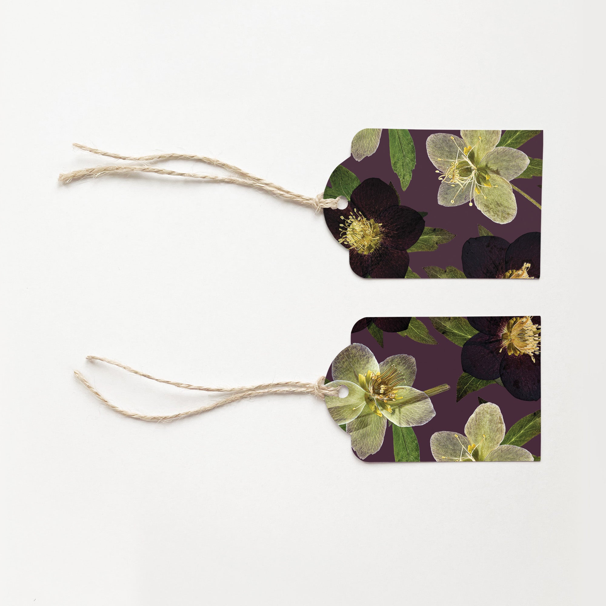 Two gift tags with natural jute hanging strings, featuring a botanical print of dark and pale hellebore 'Christmas Rose' flowers on a deep purple background, displayed on a clean white surface.