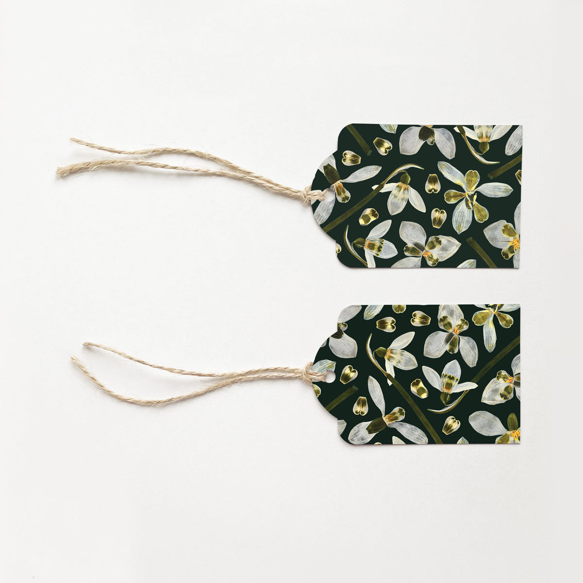 Two gift tags with natural jute string ties, featuring a dark green background printed with white snowdrop flowers, laid flat on a white background.