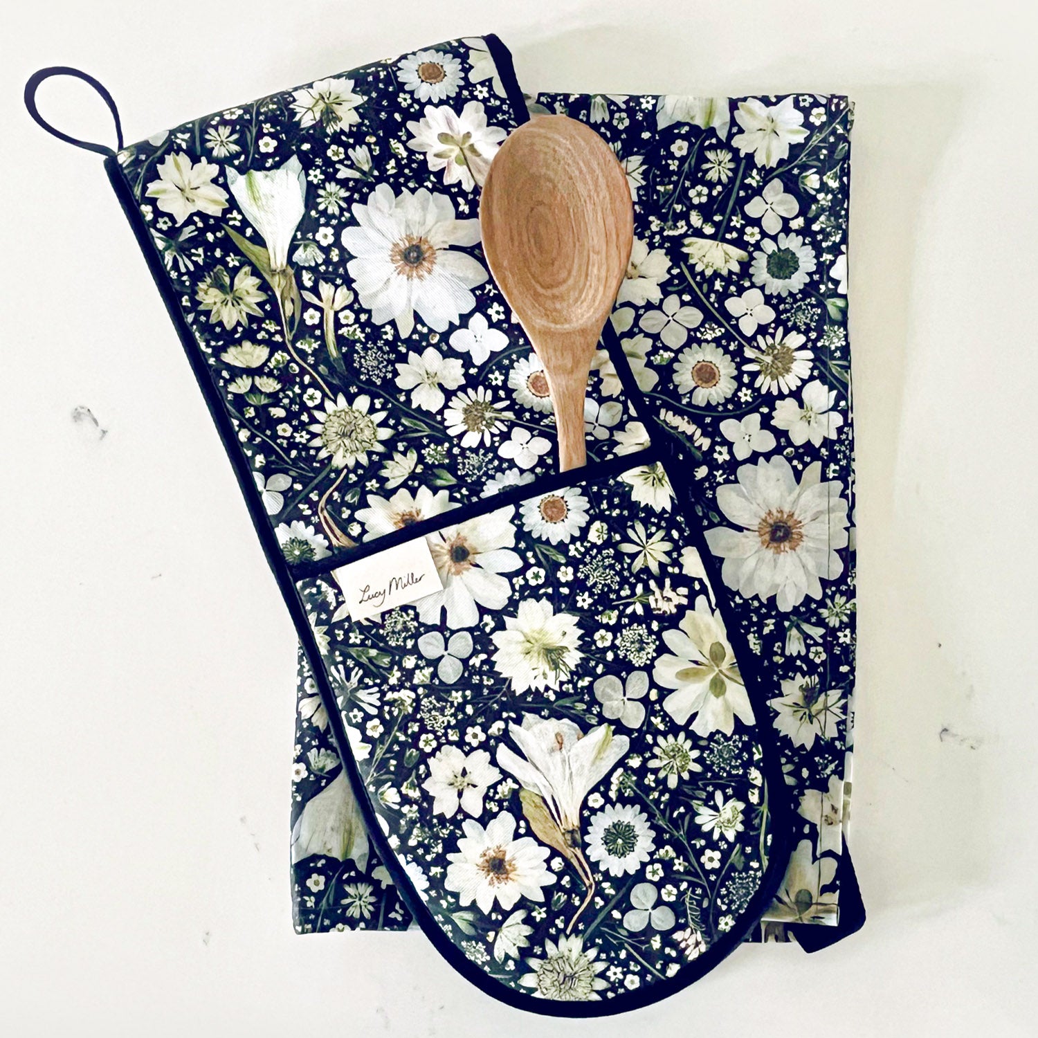 A kitchen gift set including a floral tea towel and double oven gloves. Each features a pressed flower design of white flowers on a black background. Both have a hanging loop for easy storage.