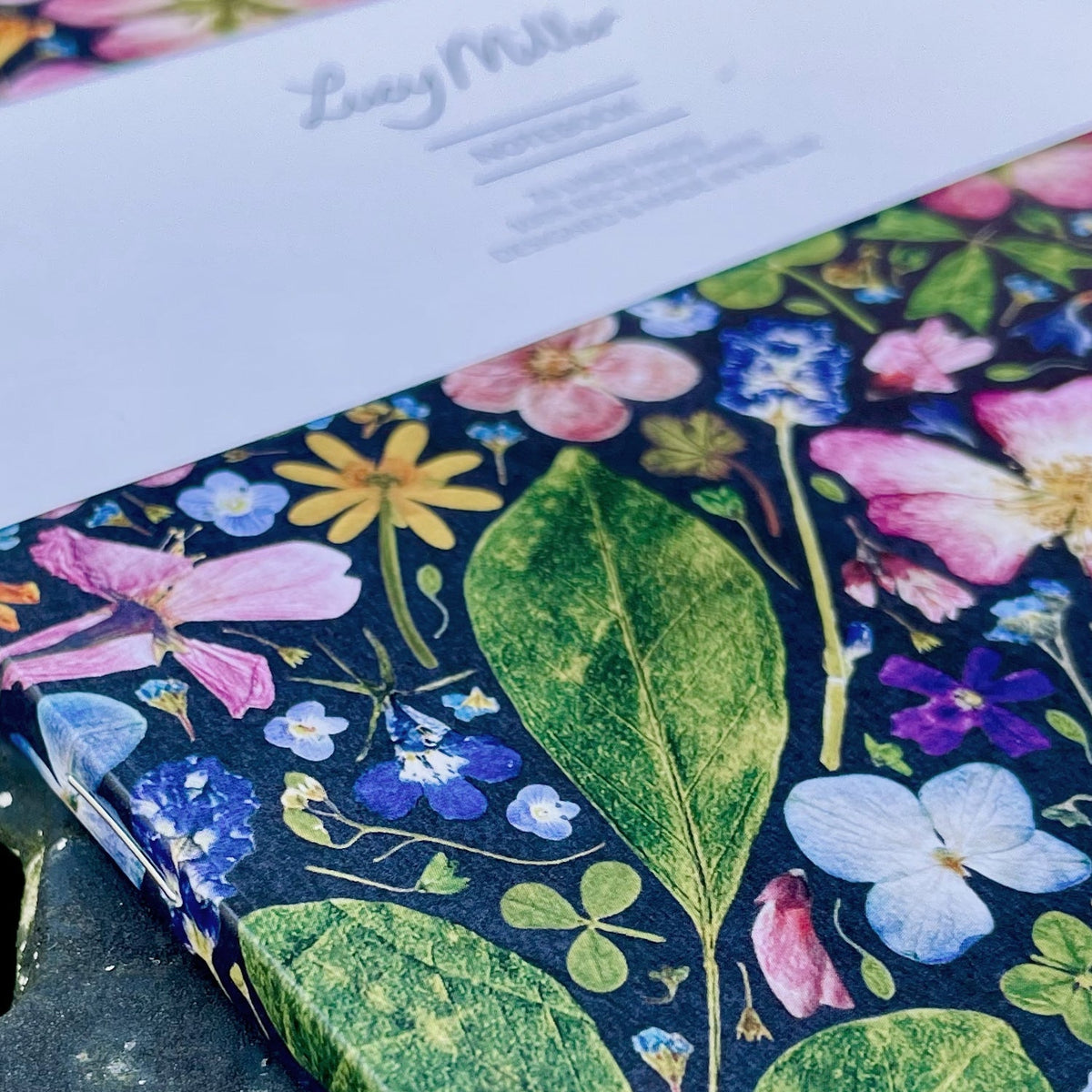 A close-up of a floral-patterned notebook or journal cover with large green leaves and pink, blue, yellow, and purple flowers on a dark background. A white paper header card with the brand name 'Lucy Miller' is partially visible at the top.