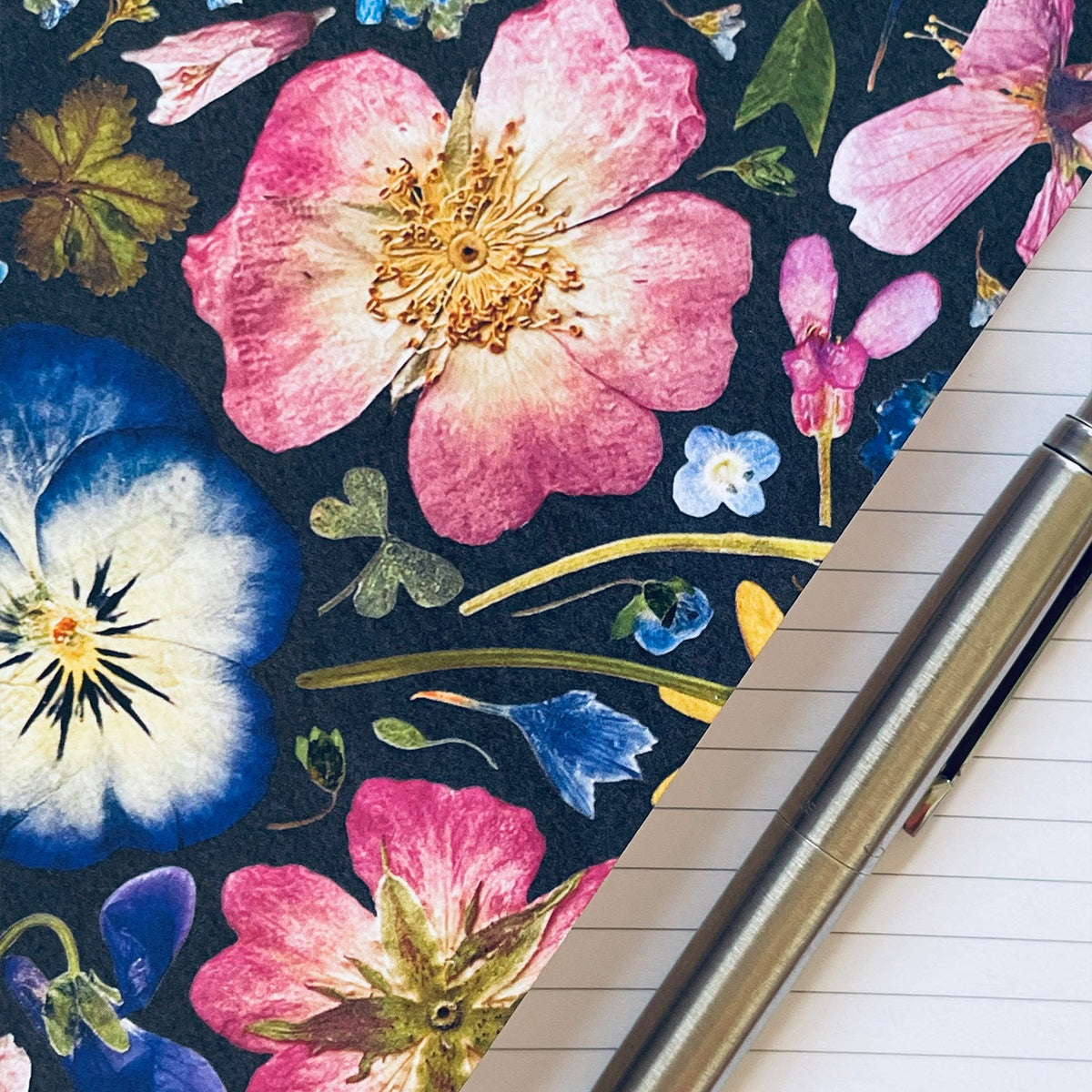 A close-up of a floral-patterned notebook cover or journal page with large pink, blue, and white flowers on a dark background, placed diagonally over a lined notebook with a metallic pen resting across it.