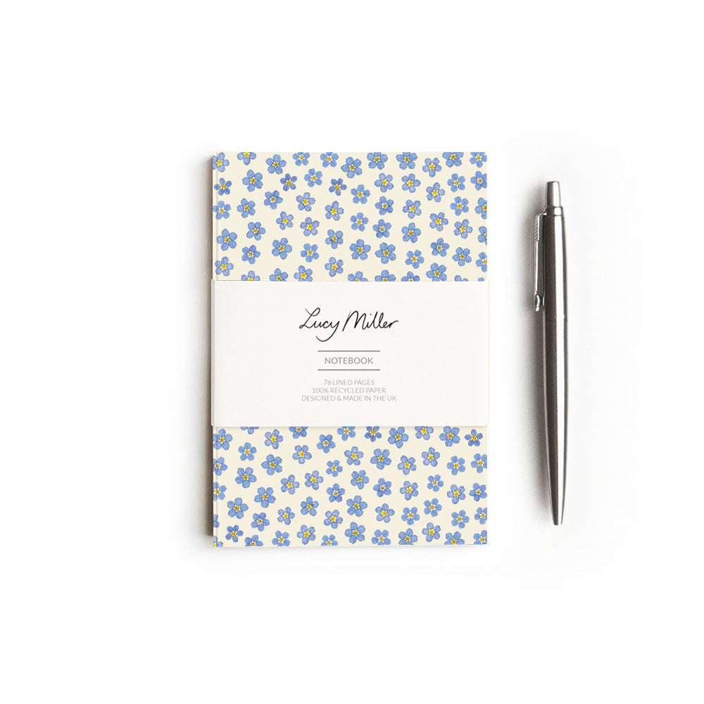 Notebook with pressed forget-me-not flowers with 'Lucy Miller' branded belly band. On a white background, and styled with a pen. 