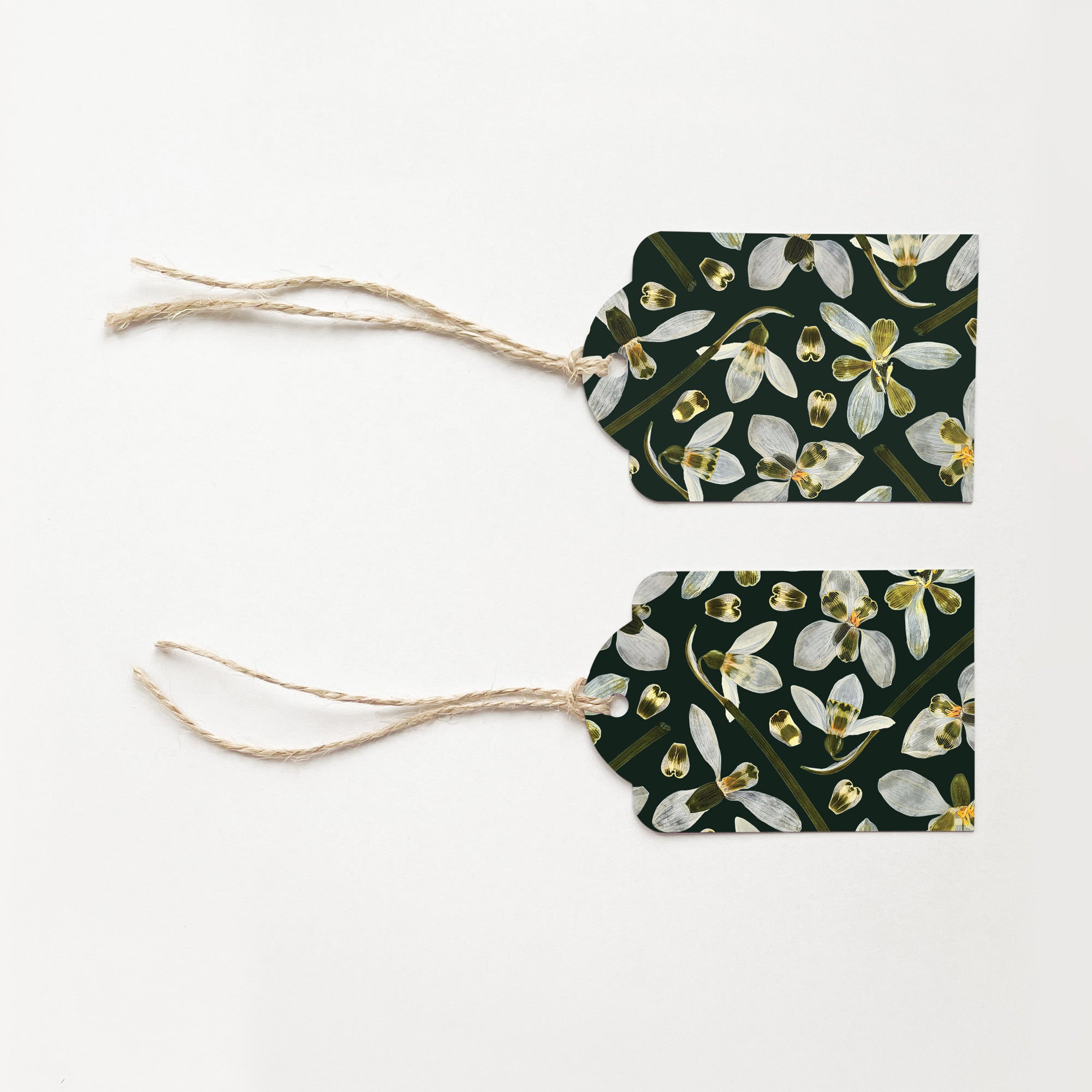 Two gift tags with natural jute string ties, featuring a dark green background printed with white snowdrop flowers, laid flat on a white background.
