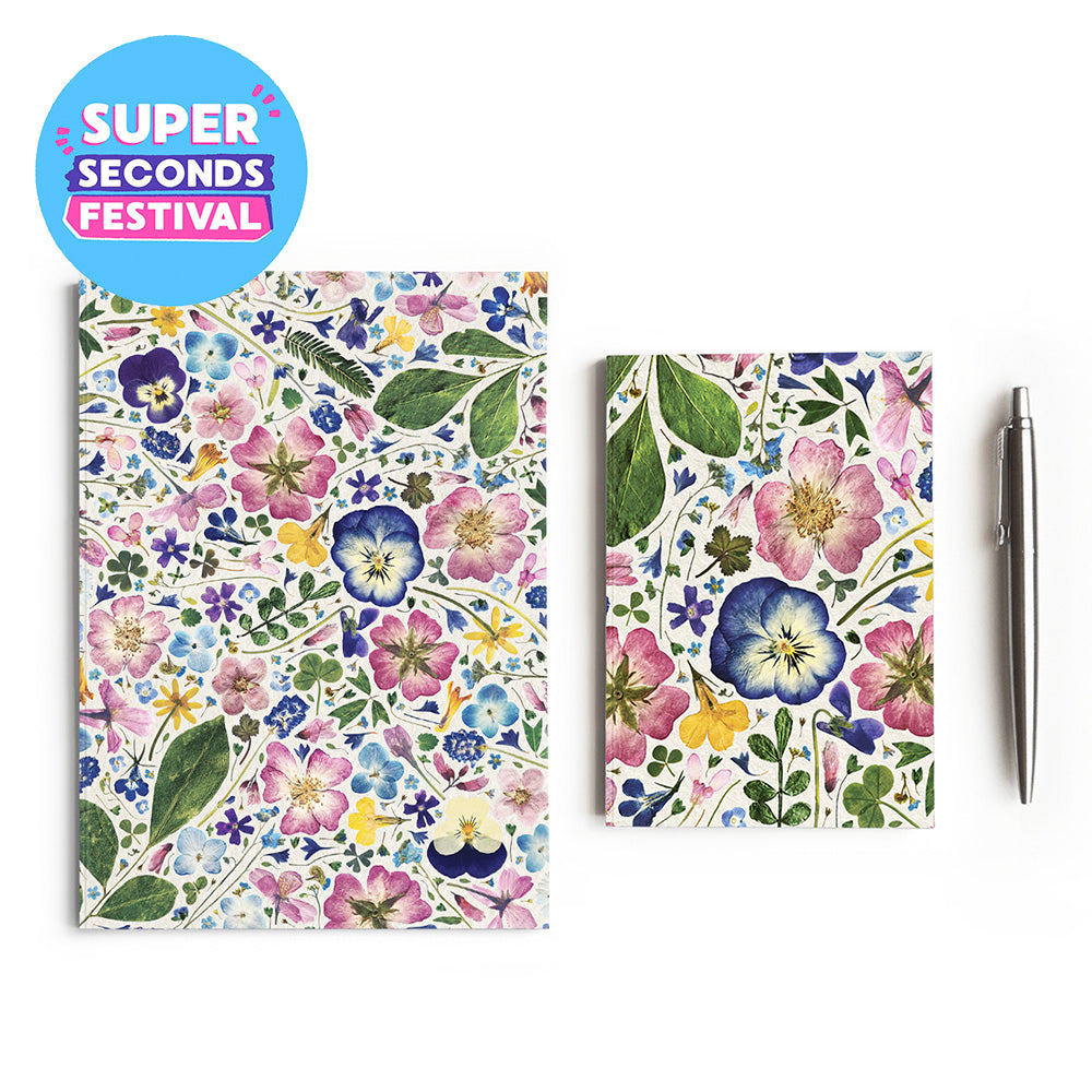 A floral-patterned notebook and matching small notebook or card set displayed on a white background, with a silver pen beside them and a blue circular badge reading “SUPER SECONDS FESTIVAL.” The floral print features pink, blue, yellow, and green flowers and leaves on a white base.
