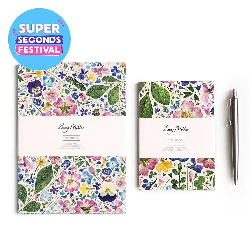 Two floral patterned notebooks with white paper bands labeled 'Lucy Miller Notebook' are shown on a white background, alongside a silver pen and a blue circular 'SUPER SECONDS FESTIVAL' badge. The notebooks feature multicolor watercolor-style flowers and leaves.