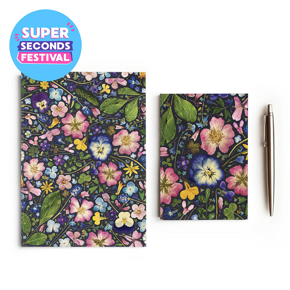 A botanical patterned notebook and matching pocket notebook displayed on a white background, alongside a metallic pen. A blue circular badge in the top-left corner reads 'SUPER SECONDS FESTIVAL'.