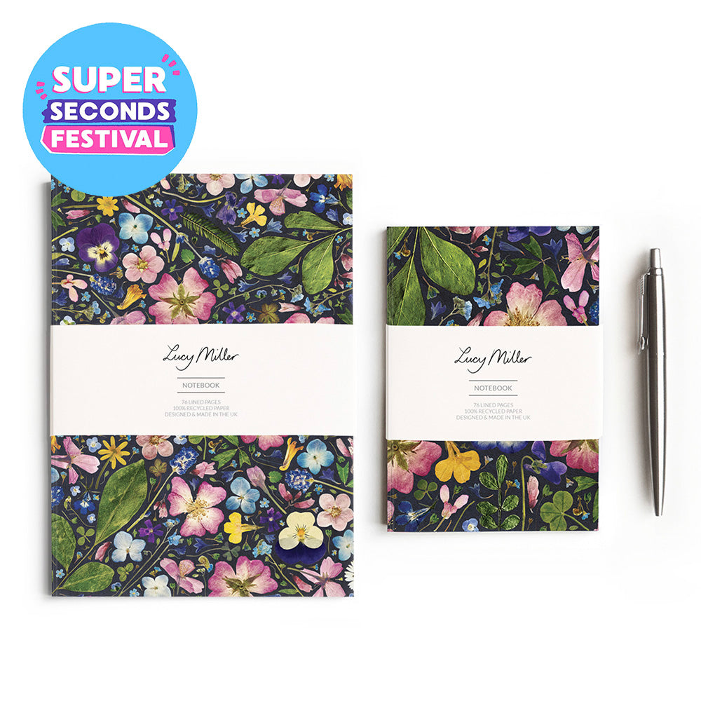 Two floral notebooks with white paper bands and a silver pen on a white background, plus a blue circular 'SUPER SECONDS FESTIVAL' badge. The notebooks show a dense multicolor botanical pattern with pink, purple, yellow, blue, and green flowers and leaves.